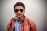 A man wearing sunglasses and a brown jacket poses casually, highlighting a modern fashion vibe.